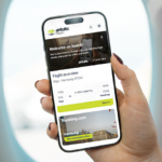 hand holding an iPhone in front of a plane window. The screen shows the new airBaltic shopping portal, including flight details and an option to book a hotel via a partner.
